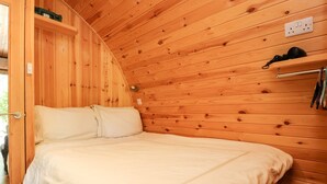 Ett sovrum i en stuga med träpanel, inrett med en säng, en hylla med en lampa och ett eluttag.