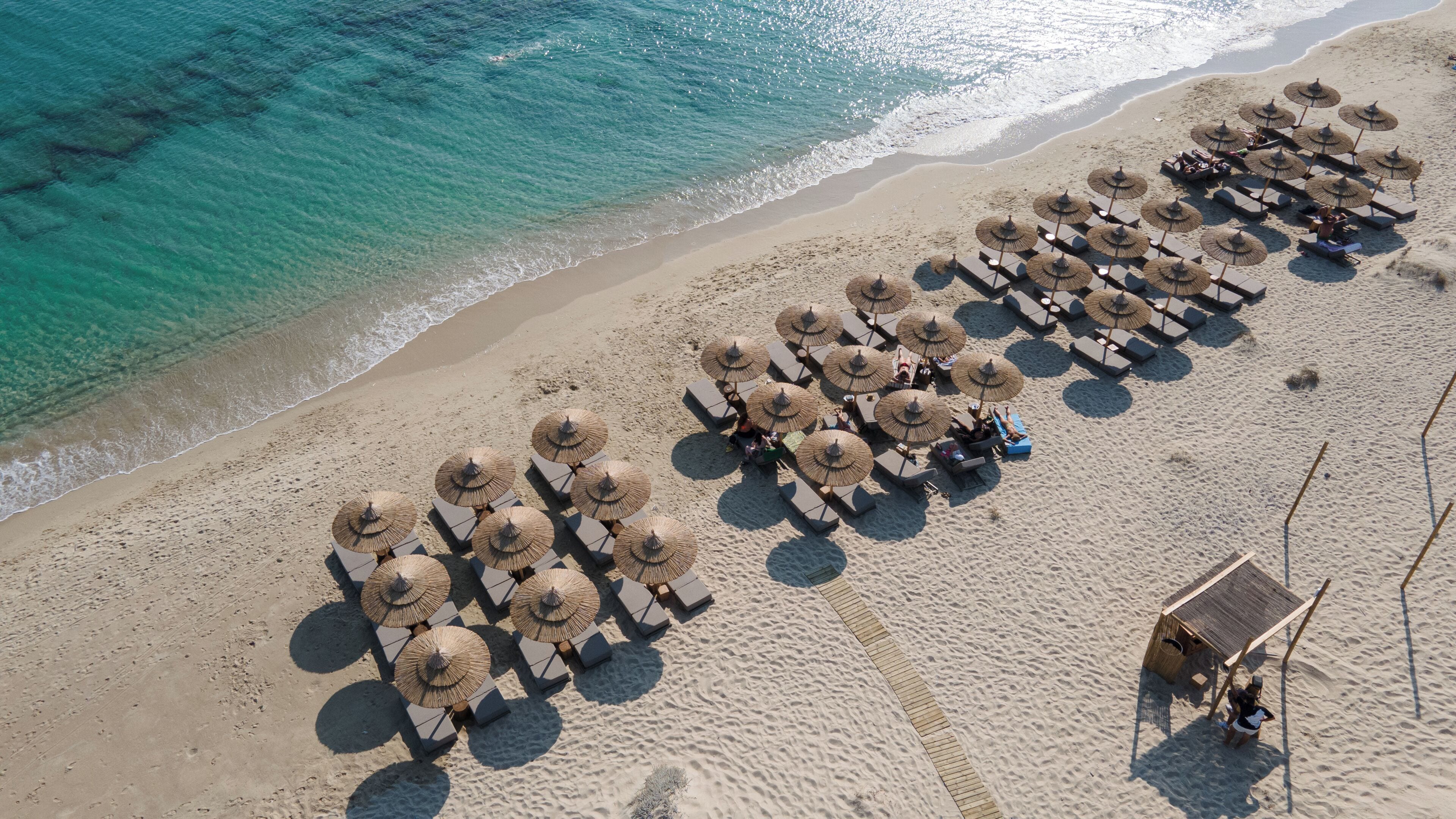Vista aérea de uma praia com fileiras de espreguiçadeiras e guarda-sóis de palha, uma pequena cabana e águas turquesa cristalinas.
