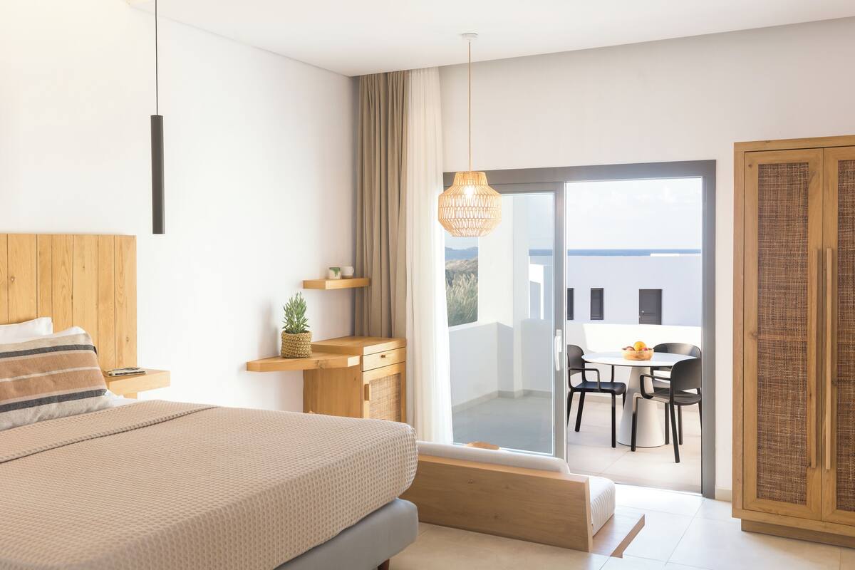 A modern bedroom with a large bed, wooden headboard, bedside table, and a view of a balcony with a dining area.