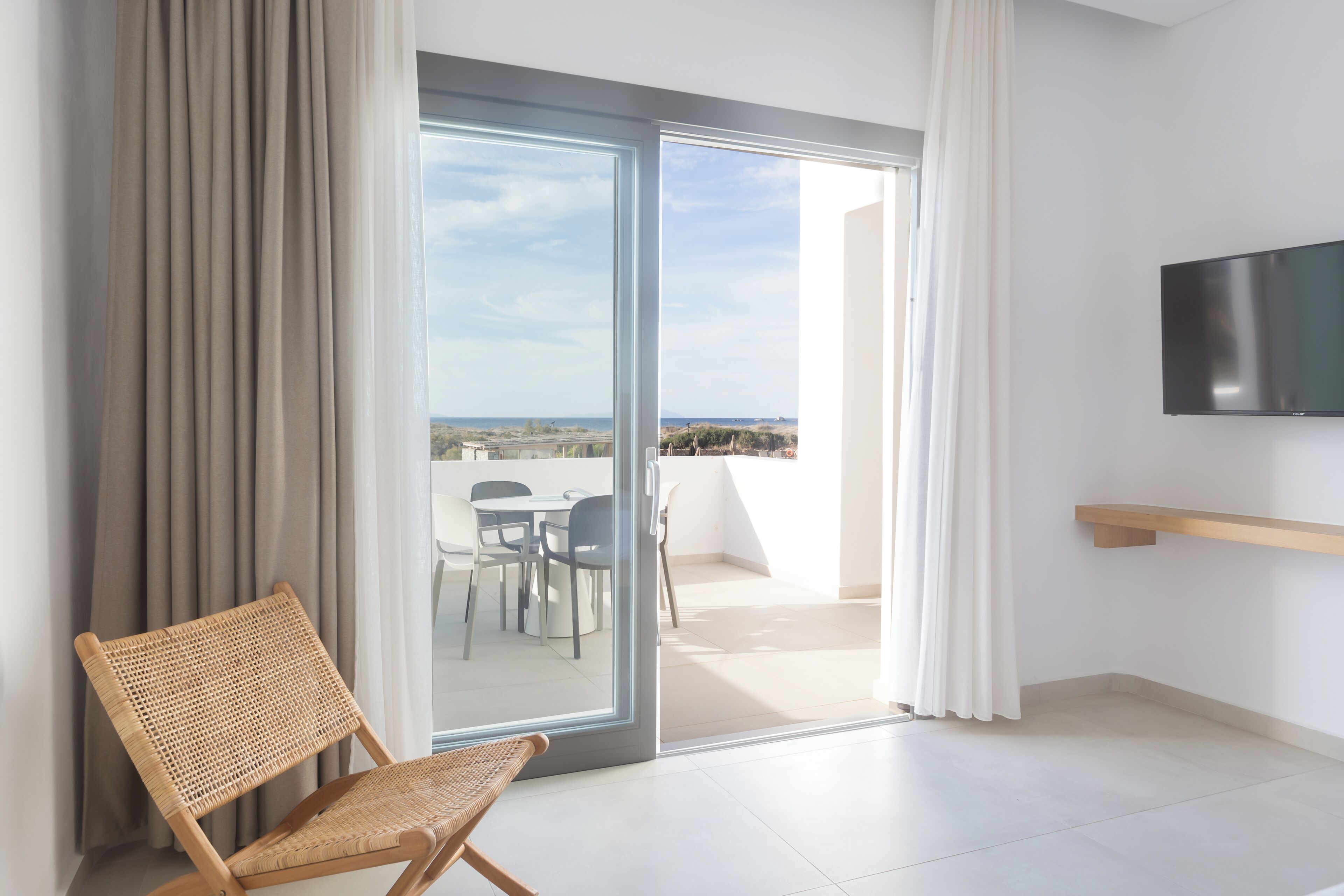 A modern room with a sliding glass door leading to a balcony, a flat-screen TV mounted on the wall, and a wicker chair.