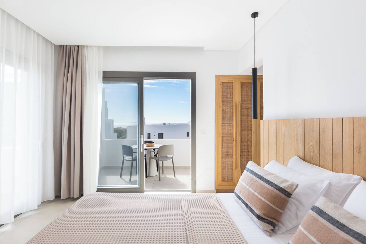 A modern hotel room with a large bed, a wooden headboard, a balcony with a table and chairs, and a view of the city.