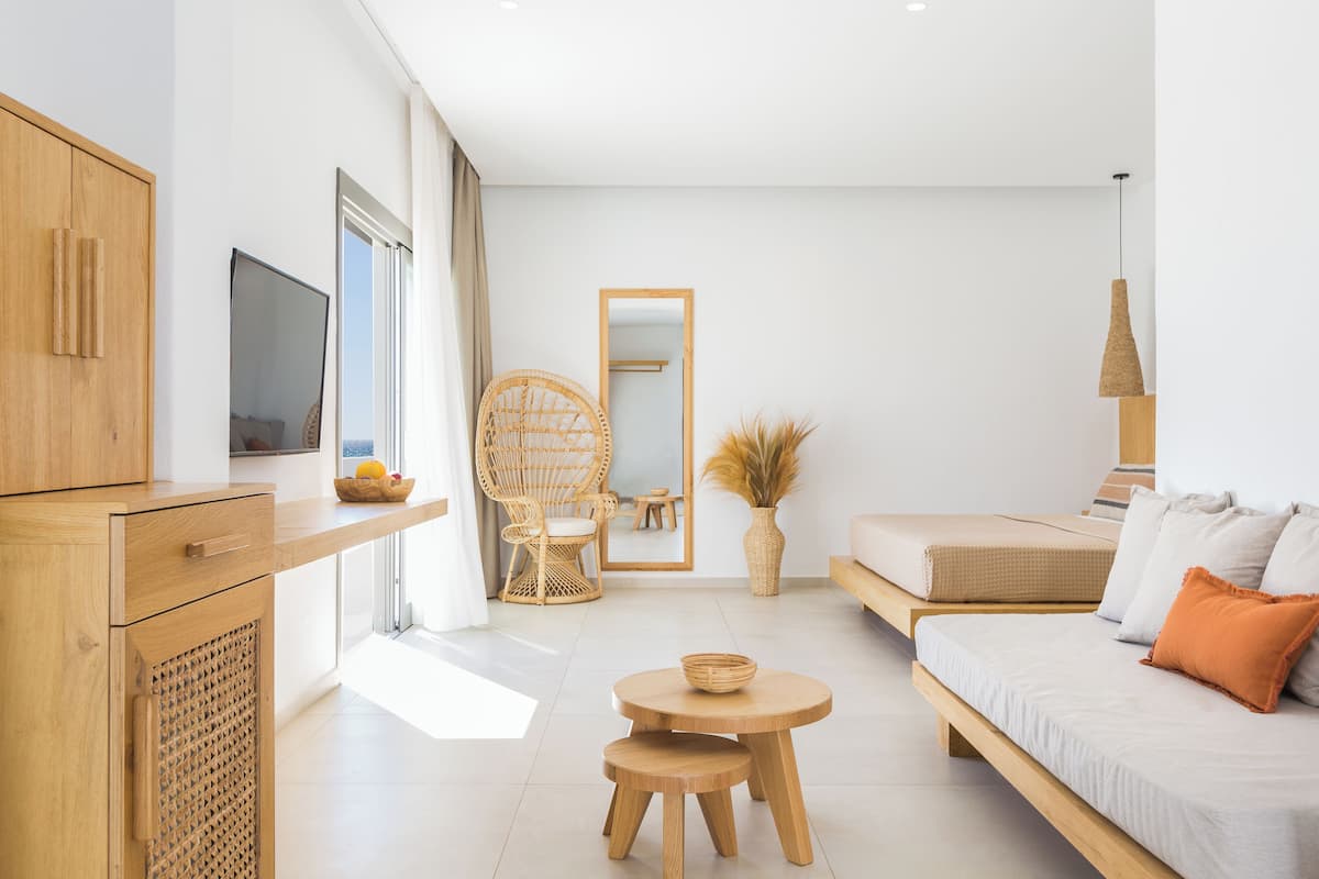 A modern, well-lit room with a wooden cabinet, a television, a wicker chair, a round table, a sofa with cushions, and a large mirror on the wall.