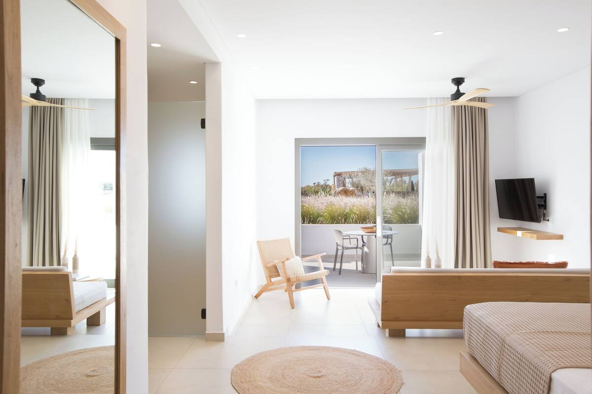A modern bedroom with a large bed, a seating area with a chair, a TV mounted on the wall, and a balcony with a view of the outdoors.