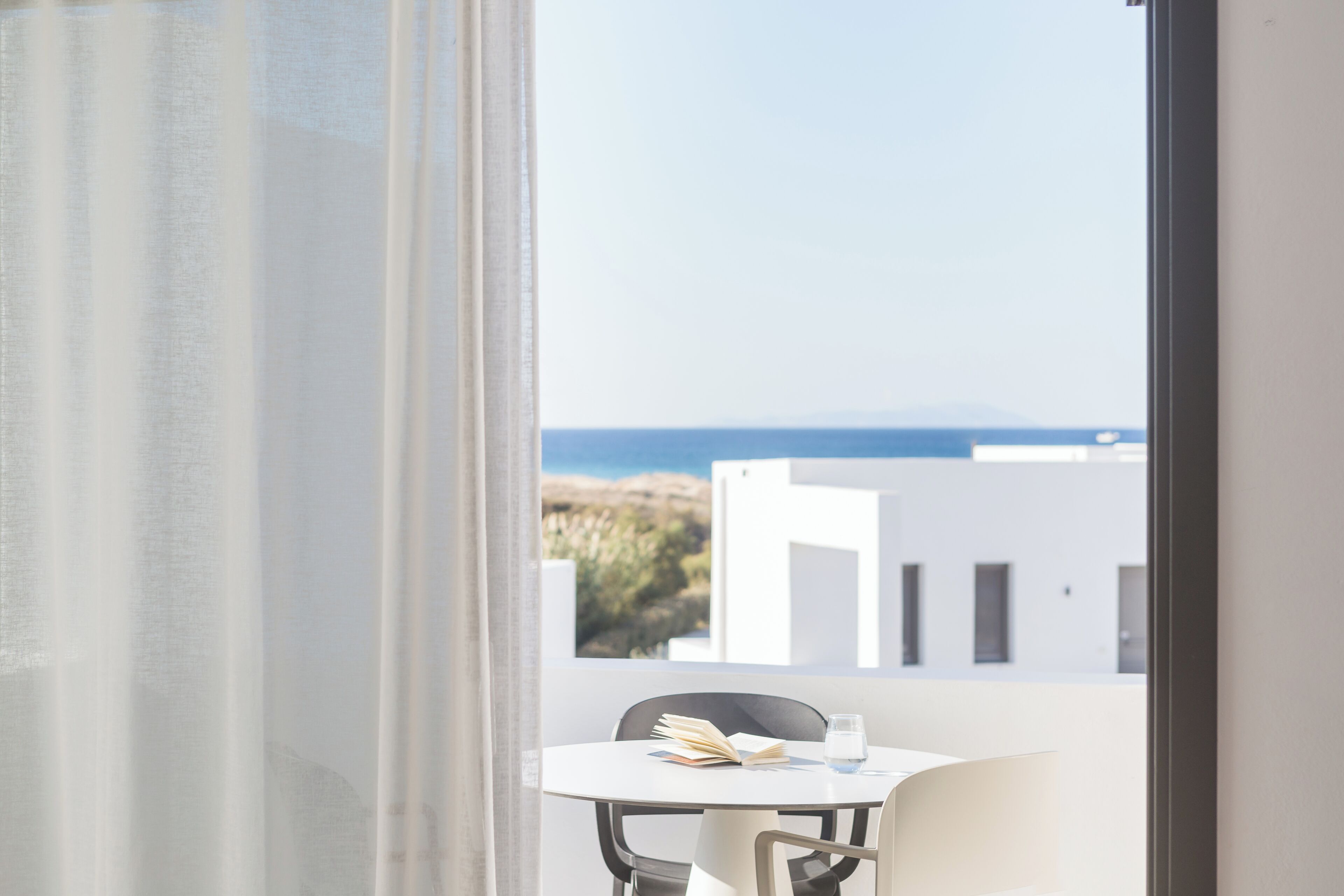 Ein Zimmer mit Blick auf das Meer und Gebäude, ein Tisch mit einem Buch und einem Glas, und ein Fenster mit transparenten Vorhängen.
