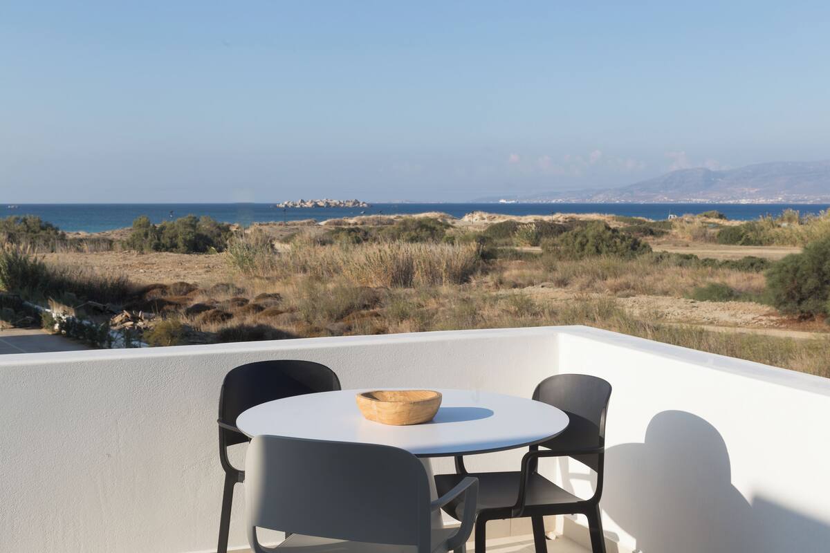 A balcony with a round table and two chairs, overlooking a coastal landscape with sparse vegetation and a distant view of the sea and city skyline.