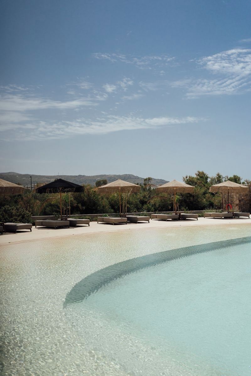 A clear pool with lounge chairs and umbrellas, surrounded by greenery and hills in the background.