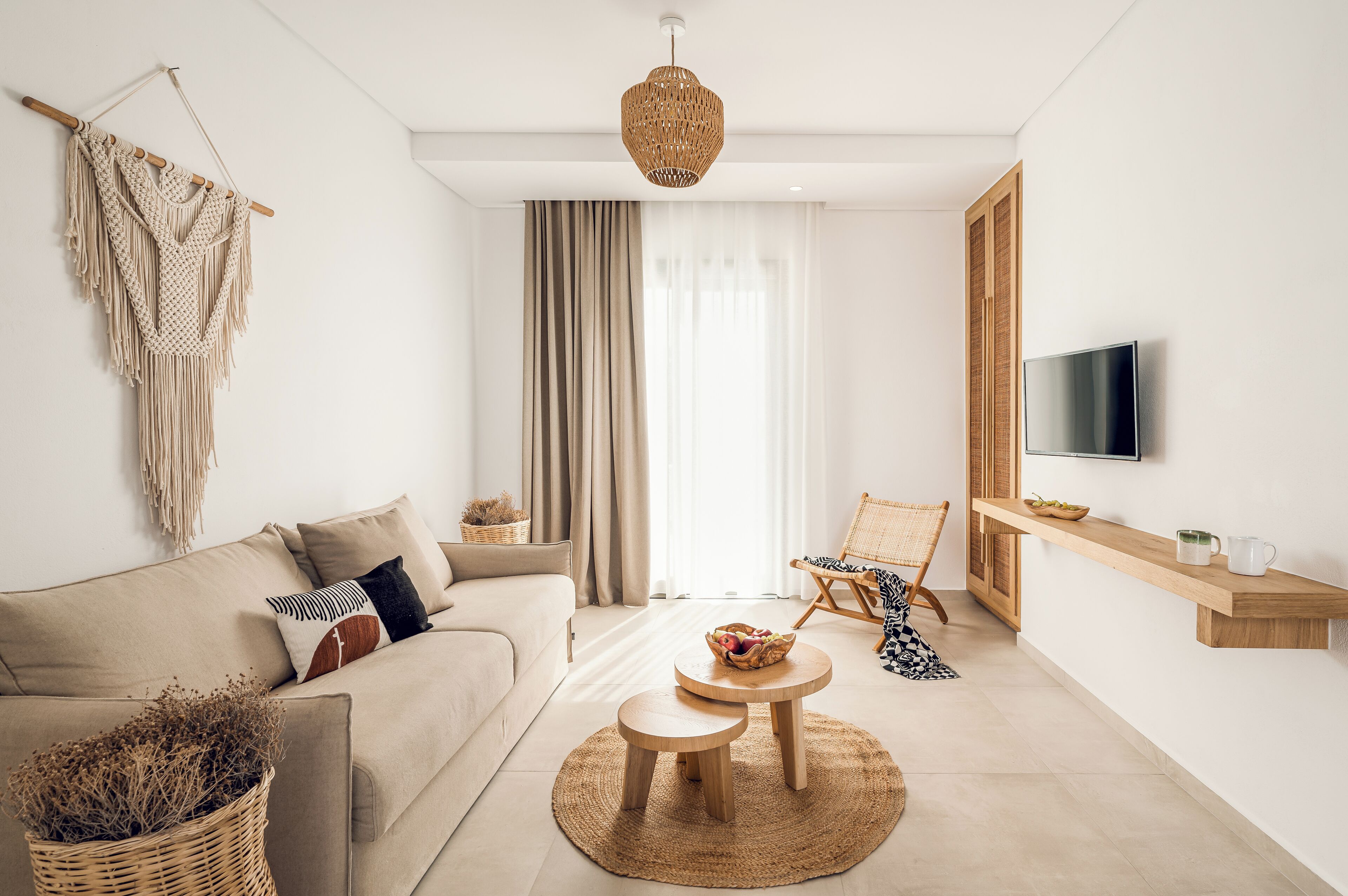 A modern living room with a sofa, two round tables, a hanging macrame wall decoration, a wicker basket, and a television on a wooden shelf.