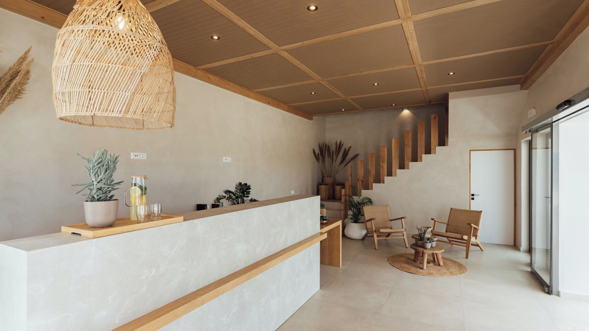 A modern reception area with a wooden reception desk, a hanging rattan light fixture, and a staircase leading to an upper level.