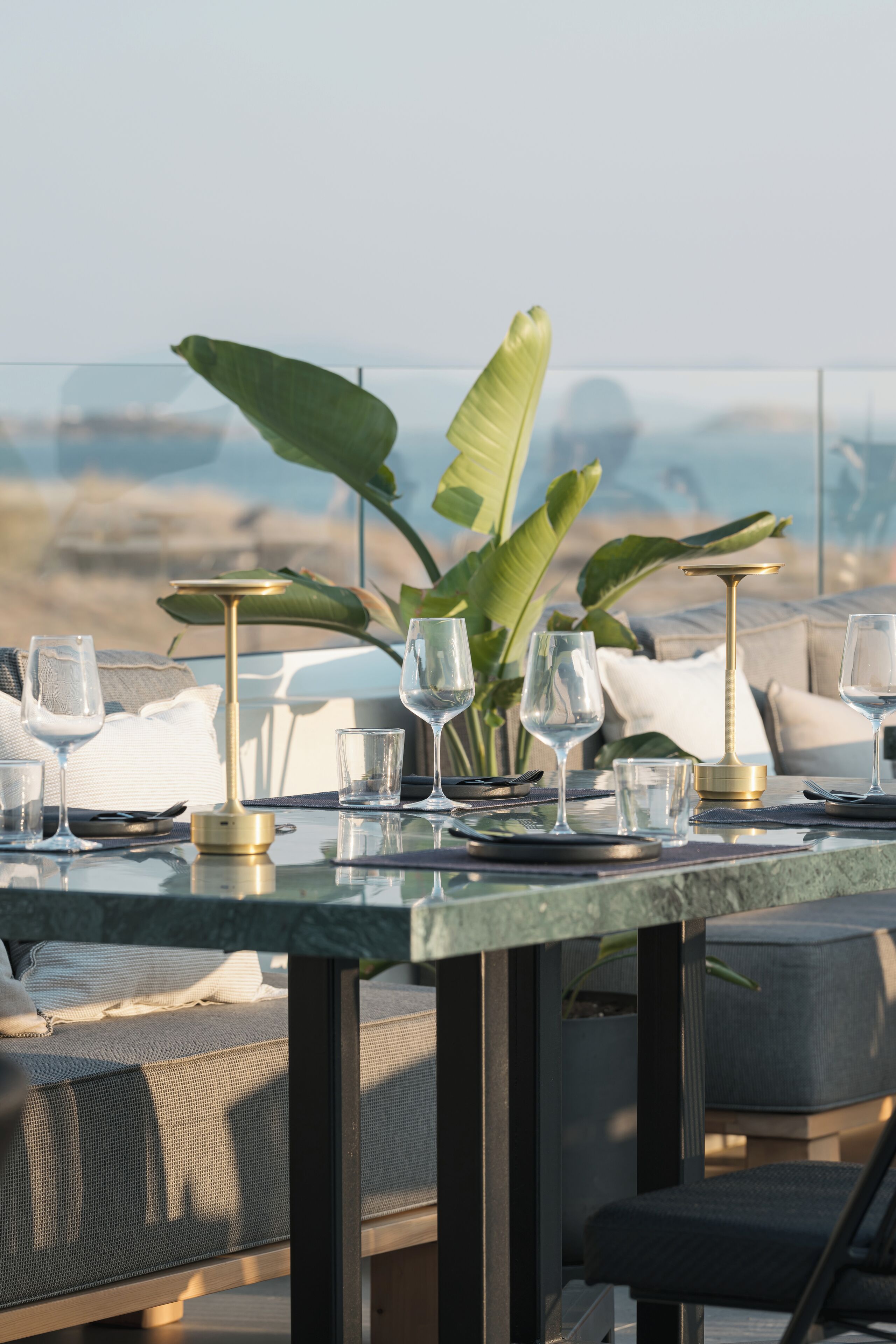 A dining table set with glasses and a gold-colored candle holder, surrounded by lush green plants and a view of the ocean.