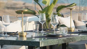 A dining table set with glasses and a gold-colored candle holder, surrounded by lush green plants and a view of the ocean.