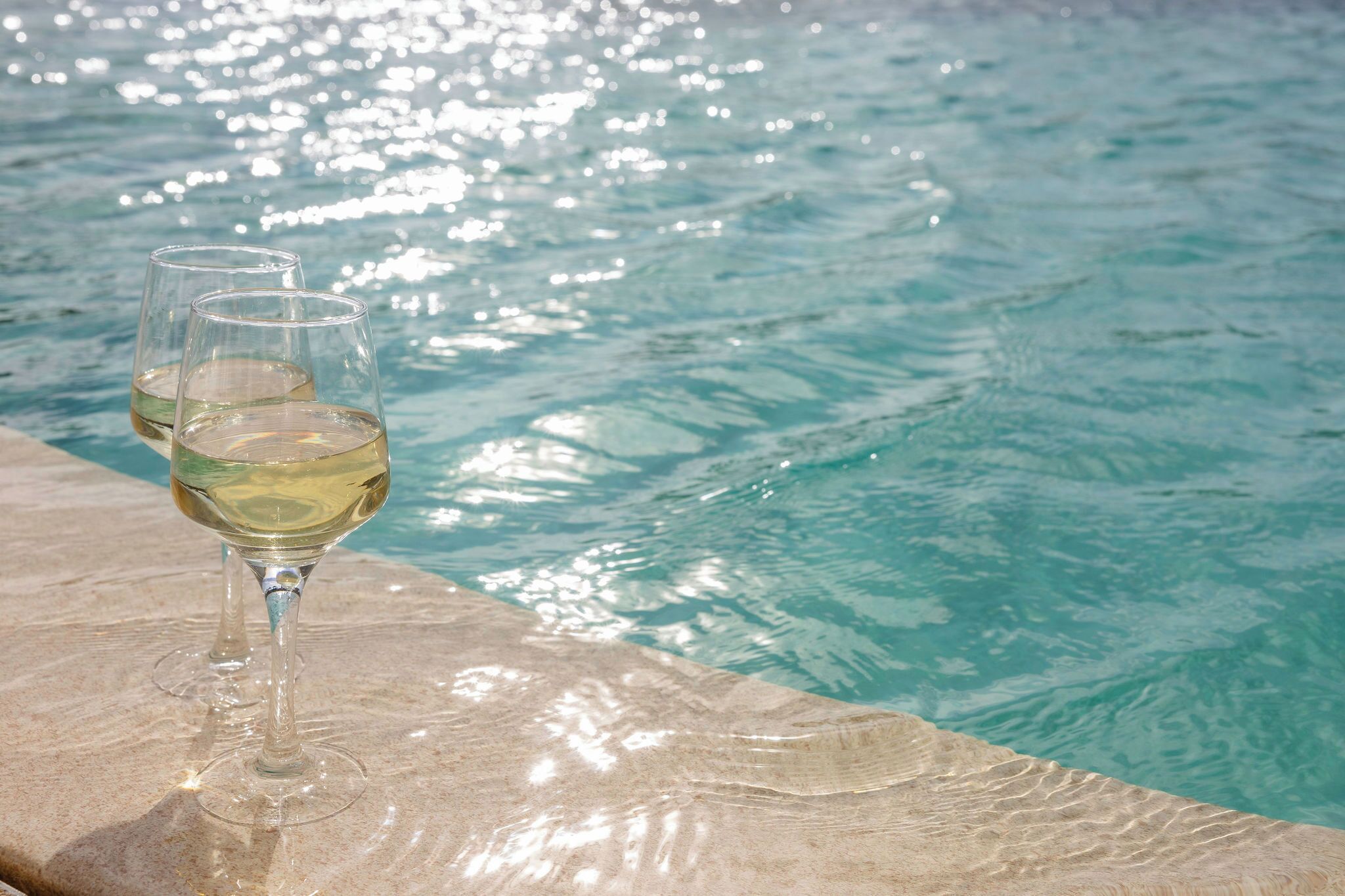 Two glasses of white wine on a stone ledge by a pool.