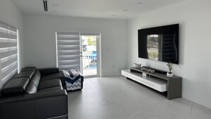 A modern living room with a black leather sofa, a flat-screen TV mounted on the wall, and a sliding glass door leading to an outdoor area. - Relaxing home in a beautiful location with luxurious accommodations (Key Largo)