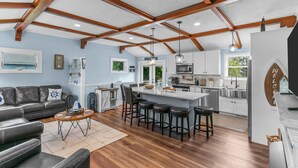 A modern kitchen with a central island, bar stools, and a dining area with a black leather couch. - Reserve Fall 2026! Beach House in the Pier W/ POOL, short walk to the beach! (Narragansett)