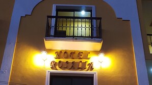 L’entrée d’un hôtel, avec une enseigne indiquant « HÔTEL ROSALIA », un balcon doté de balustrades en métal et deux appliques murales.