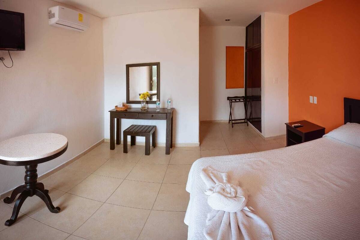 A hotel room with a bed, a desk with a chair, a small round table, a mirror, and an air conditioning unit.
