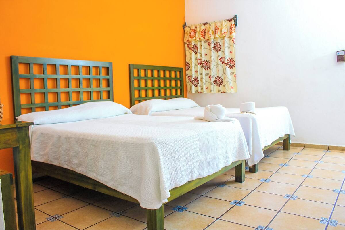 A room with two beds, each with a wooden headboard, white linens, and a small table with a cup and saucer.