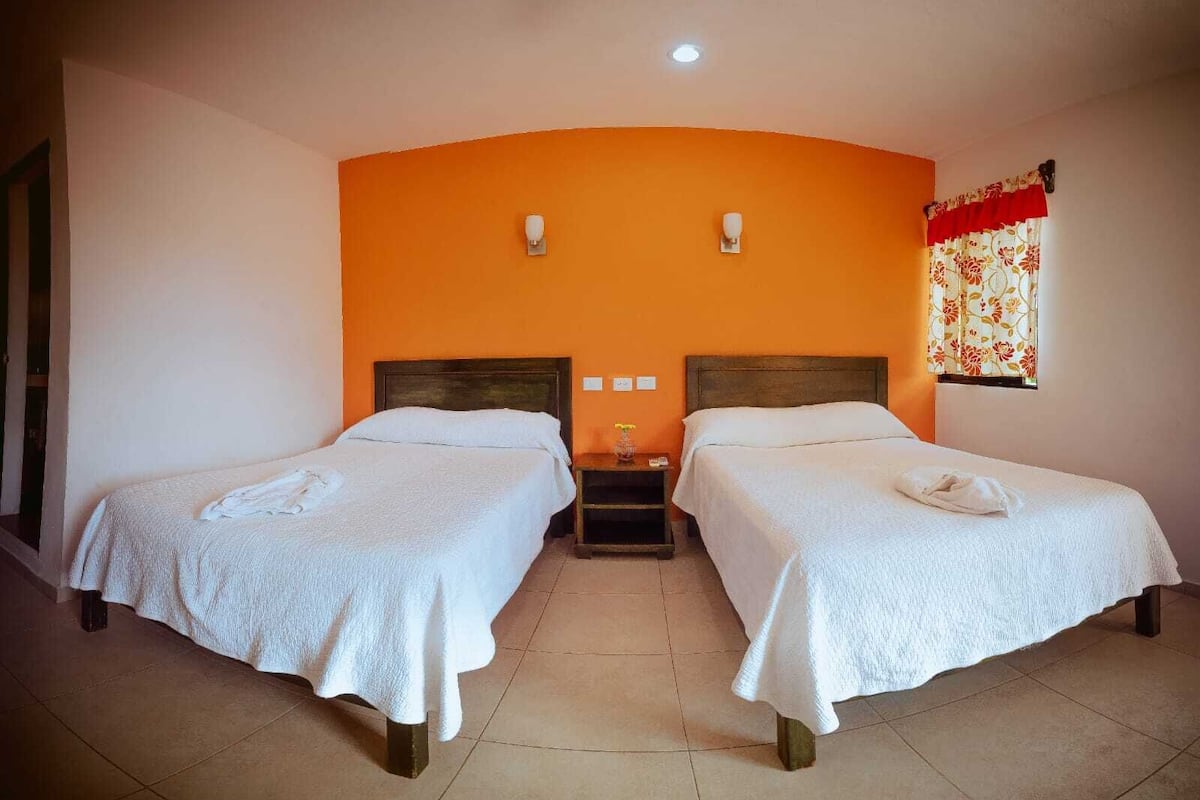 Una habitación con dos camas, una mesita de noche, una lámpara de pared y un ventanal con cortinas.