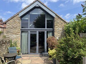 A stone house with a large glass door and windows, a patio with a table and chairs, and a garden with various plants. - The Nail Shed (Scarborough)