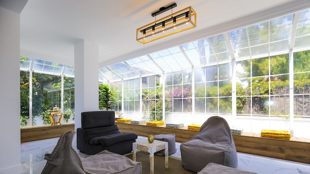 A conservatory with a glass ceiling, seating area with chairs and a table, and a view of greenery outside.