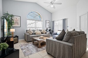 A modern living room with a grey sofa set, a coffee table, a TV, and a ceiling fan. - 618 - Bright 4 Bedroom Villa w/ Private SE Pool Near Disney (Davenport)
