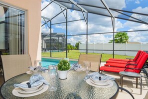 Un espacio comedor al aire libre techado, con una mesa de vidrio, sillas rojas y vista a una piscina.