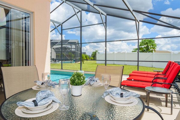A covered outdoor dining area with a glass table, red chairs, and a view of a swimming pool.