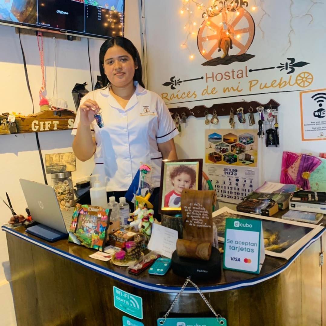 A hotel reception desk with a sign reading "Hostal Raíces de mi Pueblo," a laptop, a gift bag, and a "Se aceptan tarjetas VISA" sign.