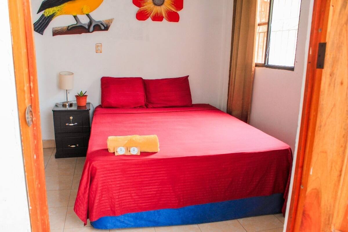 Une chambre à coucher avec une couvre-lit rouge, deux oreillers, une petite table de chevet avec une lampe et une plante, et une décoration murale représentant un oiseau et une fleur.