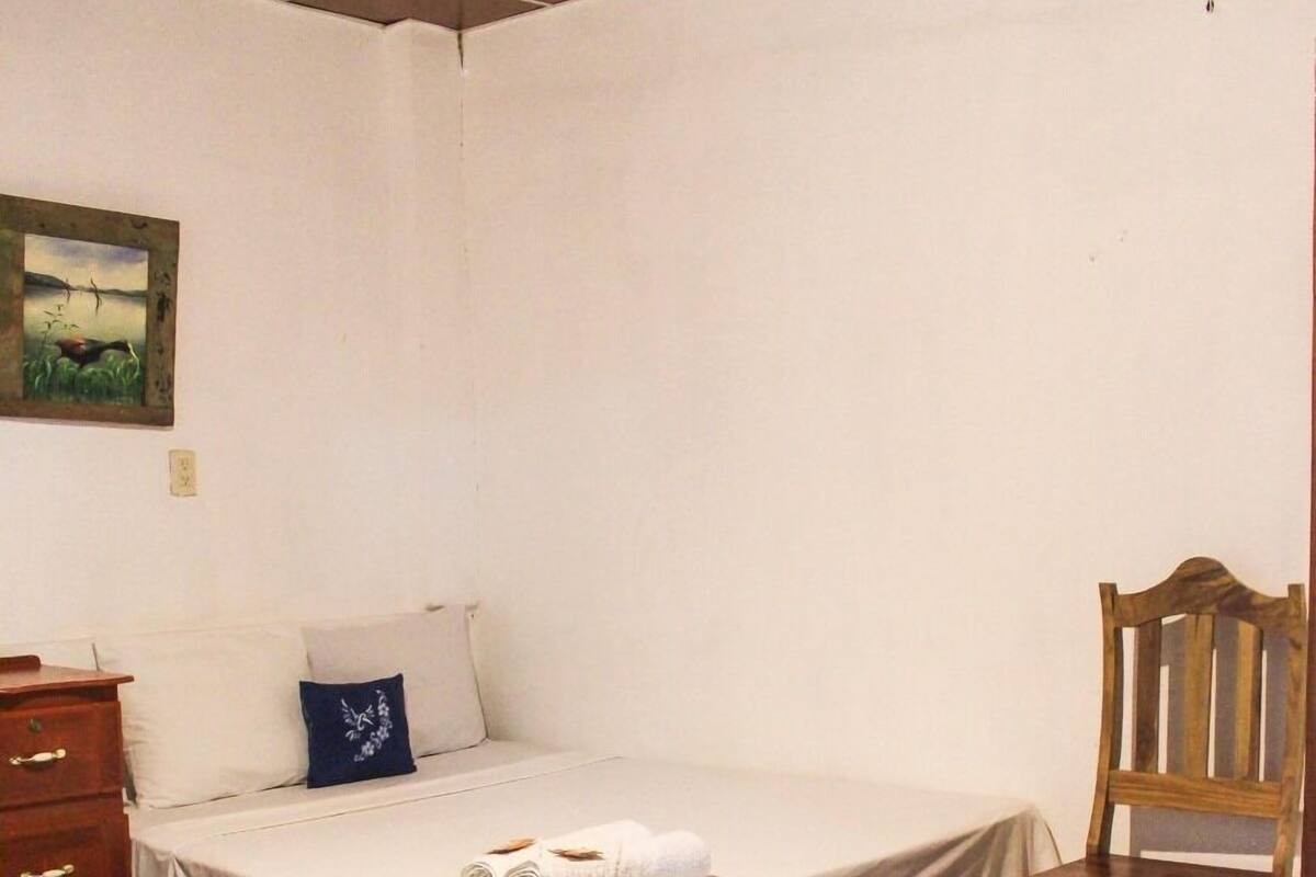 Une chambre à coucher avec un lit, une chaise en bois, une commode et un tableau accroché au mur.