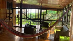 Une pièce avec un hamac, des fauteuils en osier et une vue sur la verdure grâce à de grandes fenêtres.