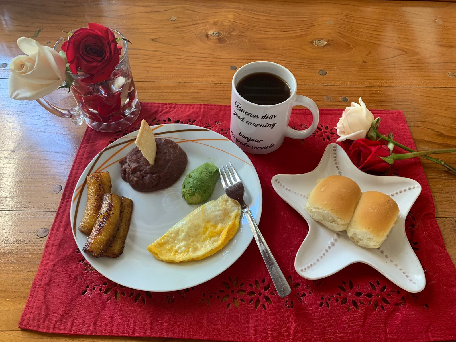 A breakfast plate with eggs, beans, avocado, and plantains, a coffee mug, a glass of water with a rose, and a star-shaped plate with buns.