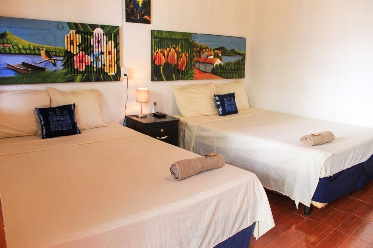 Une chambre d’hôtel avec deux lits, chacun recouvert d’une courtepointe blanche et agrémenté d’oreillers bleus. Des tableaux représentant des scènes de nature ornent les murs.