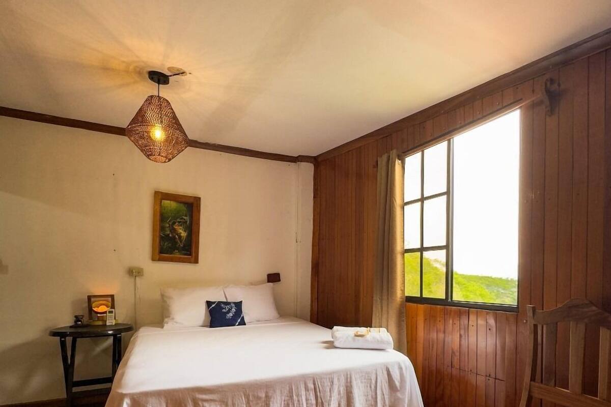 Une chambre à coucher avec un lit en bois, une table de chevet, un banc et une grande fenêtre donnant sur un paysage verdoyant.