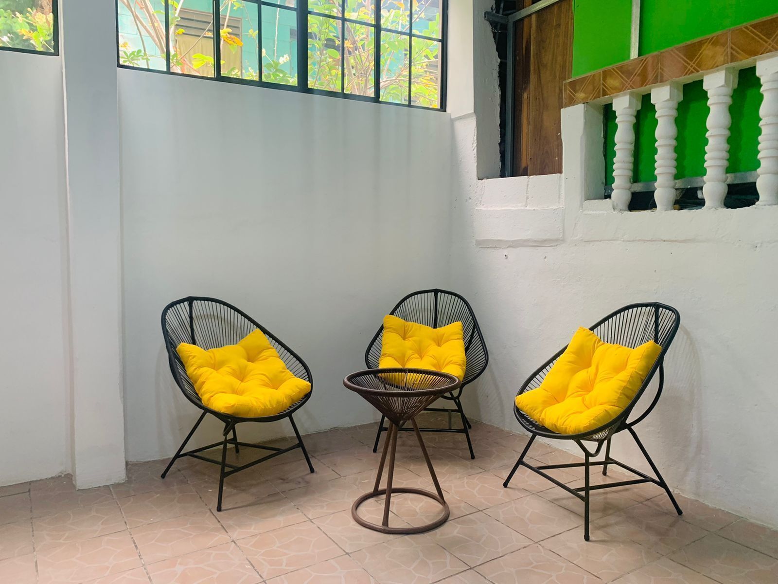 A corner room with three black wireframe chairs with yellow cushions, a small round table, and a view through a window with a green wall and white columns.