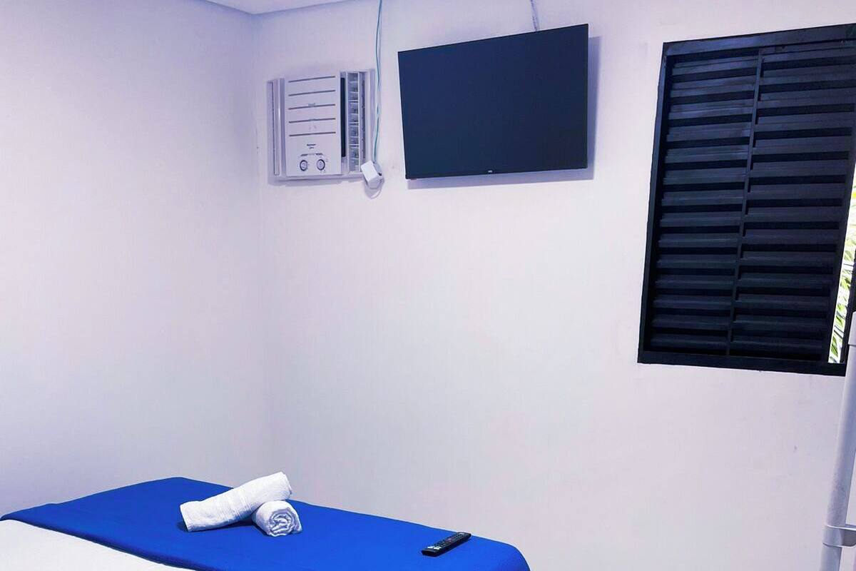 A single bed with a blue bedspread, a white pillow, and a rolled towel. There is a remote control on the bed. The room has white walls, a black window shutter, and a white air conditioning unit on the wall.