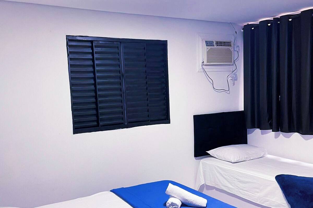 A hotel room with two single beds, a flat-screen TV, an air conditioning unit, and a window with black shutters.