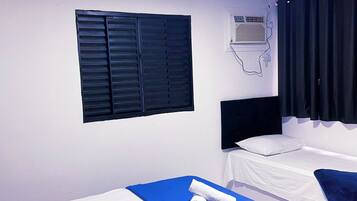 A hotel room with two single beds, a flat-screen TV, an air conditioning unit, and a window with black shutters.
