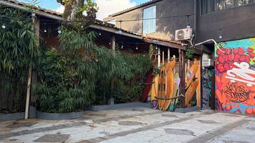An outdoor area with a tiled floor, a tree, and surfboards leaning against a wall.