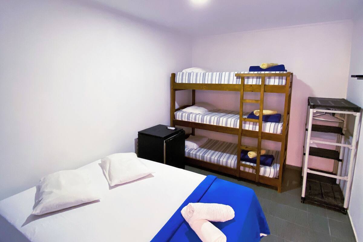 A single bed with a blue bedspread and white pillows, a bunk bed with striped bedding, a black nightstand, and a white shelf unit.