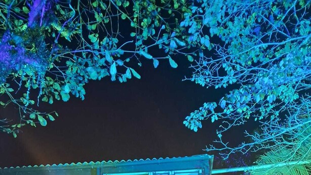 Ένας εξωτερικός χώρος για φαγητό με τραπέζια και καρέκλες, ένα κτίριο με κεκλιμένη στέγη και φωτιστικά που κρέμονται σε σειρές.