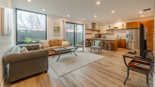 Amplia sala de estar con sofá, mesa de centro y sillas, cocina con electrodomésticos modernos y ventanas grandes con vistas.