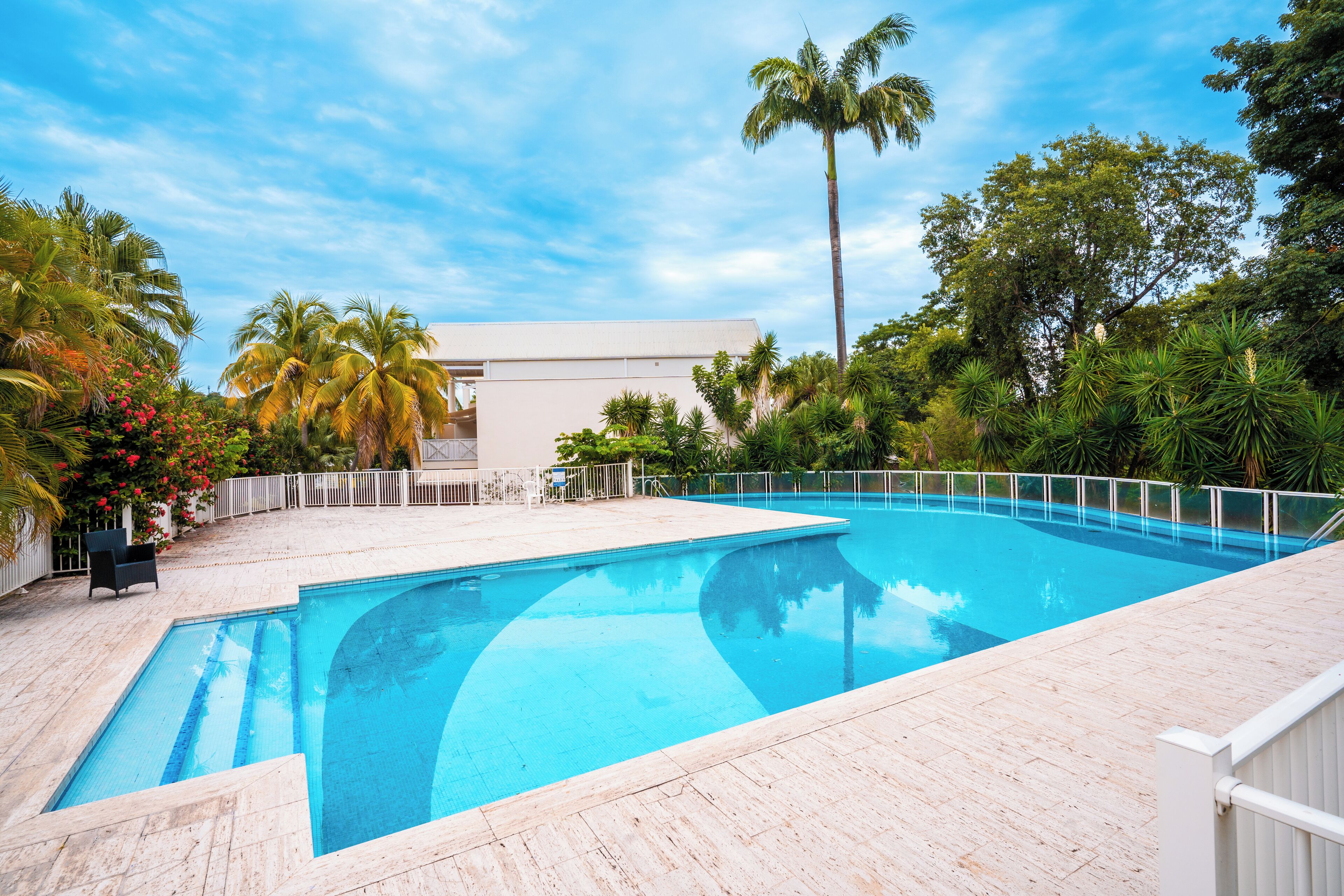 Ein rechteckiger Swimmingpool mit einer weißen Poolterrasse, umgeben von Palmen und tropischer Bepflanzung.