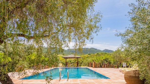 Piscina rectangular con una pérgola y sillones de descanso en un jardín ajardinado con vistas a colinas distantes.