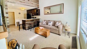 Ein modernes Wohnzimmer mit einer Holzwand, einer Küchenzeile mit dunklen Schränken, einem beigen Sofa mit gemusterten Kissen und einem großen Spiegel an der Wand.