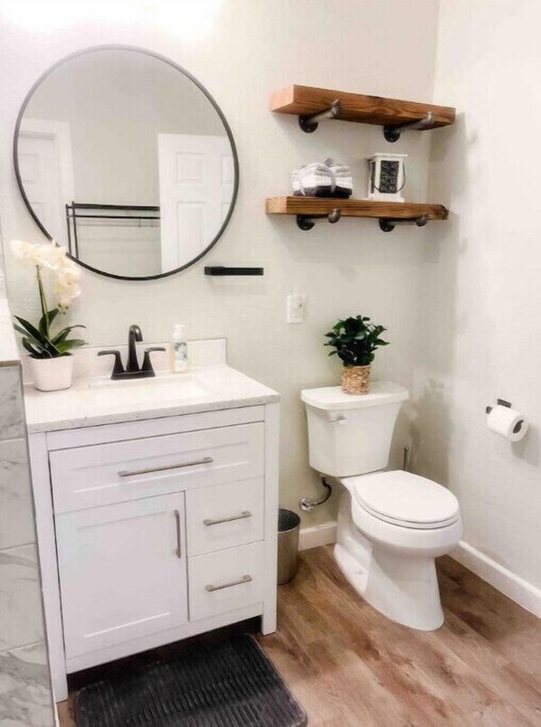A bathroom with a white vanity, a round mirror, a toilet, and wooden shelves above the toilet. - Lakeview Oasis (Nevada)