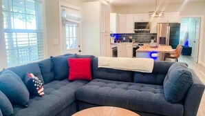A modern living room with a dark blue sectional sofa, a round wooden coffee table, and a kitchen in the background with white cabinets and stainless steel appliances. - Lakeview Oasis (Nevada)