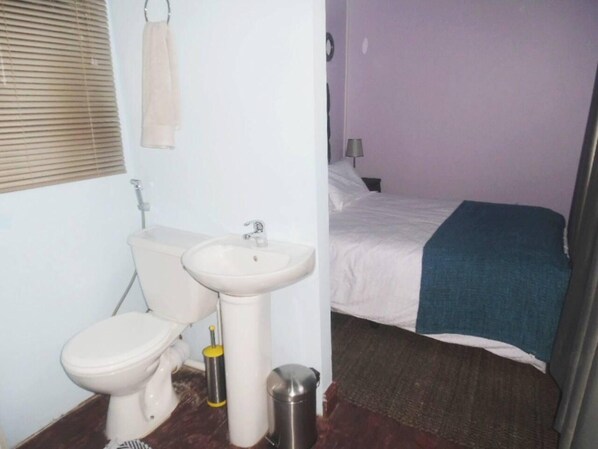 Un baño con inodoro, lavamanos y bidé. Se ve una cama con una manta azul y una lámpara en una mesita de noche.