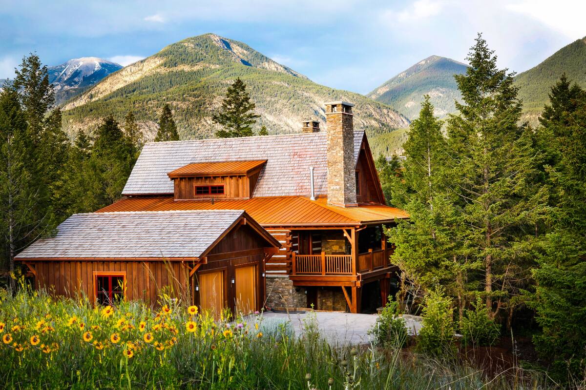 A log cabin with a stone chimney, wooden deck, and a mountain backdrop.