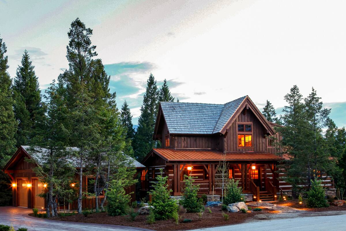 A large log cabin with a prominent front porch and multiple windows, surrounded by dense forest.