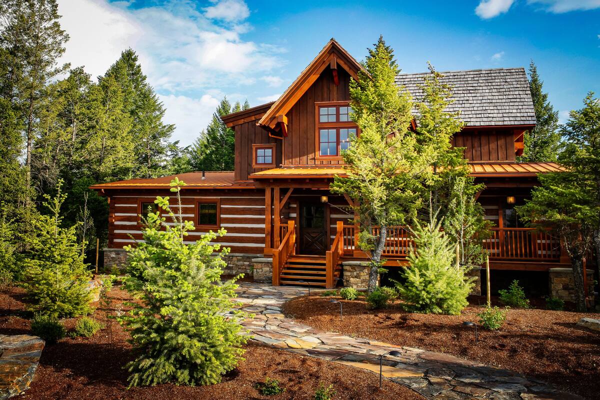 A log cabin with a stone pathway leading to the entrance, surrounded by trees and a clear sky.